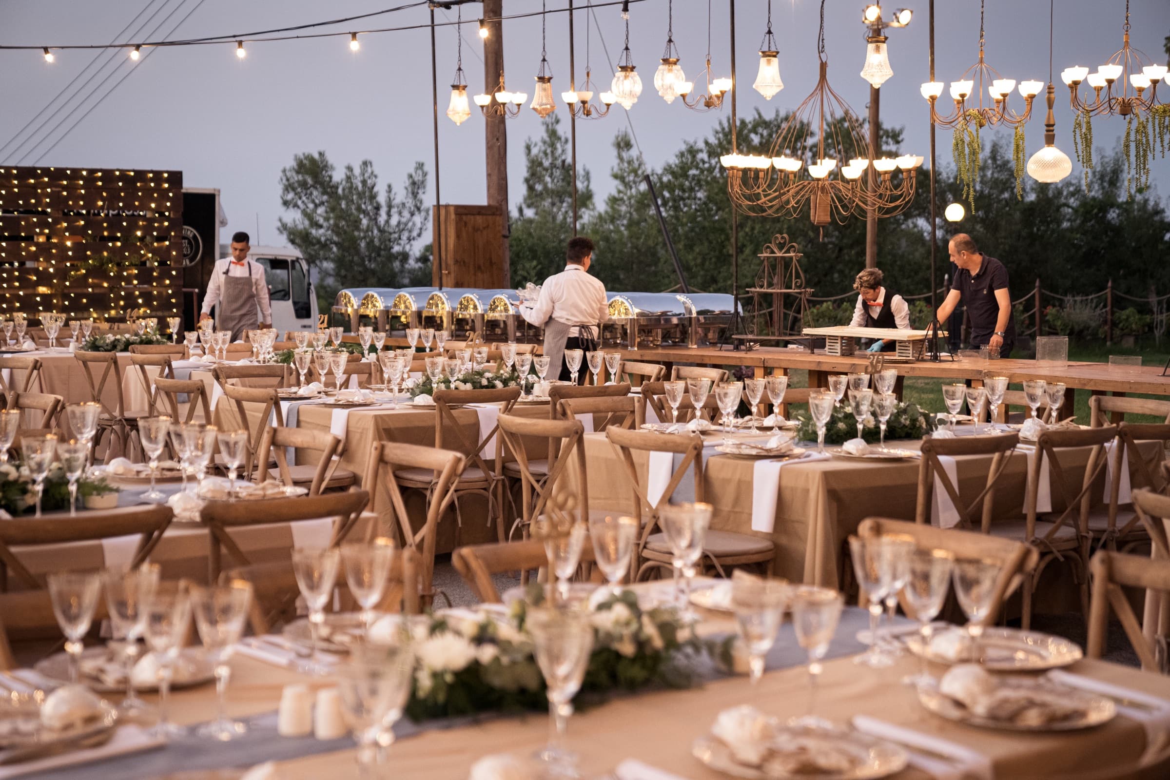 The long table set under the pines at dusk, chandeliers lit
